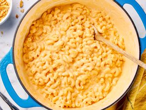 One pot mac and cheese in a dutch oven with a wooden spoon, and in the surroundings, a bowl with cracker topping and a mustard kitchen towel