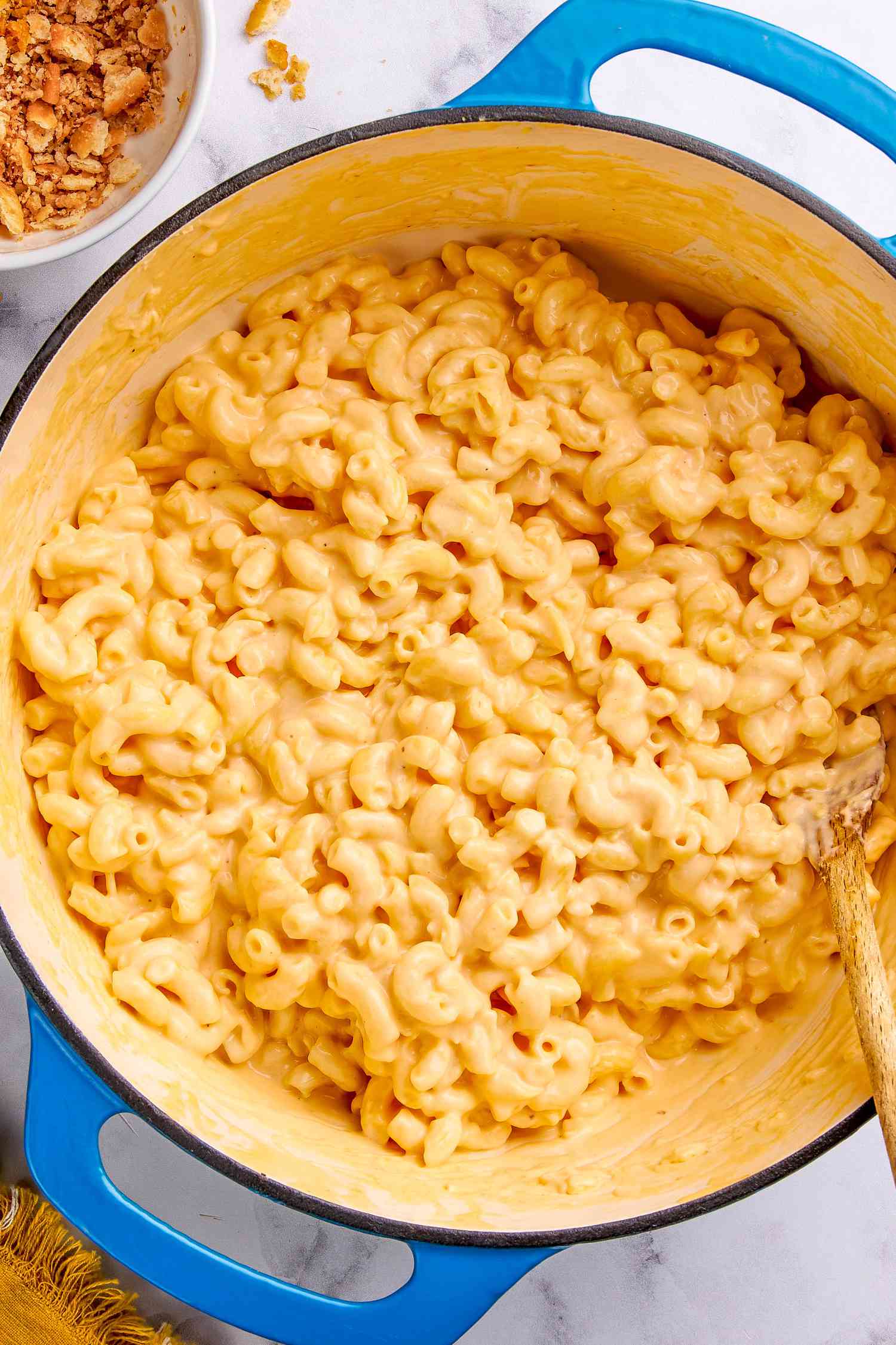 One pot mac and cheese in a dutch oven with a wooden spoon, and in the surroundings, a bowl with cracker topping and a mustard kitchen towel