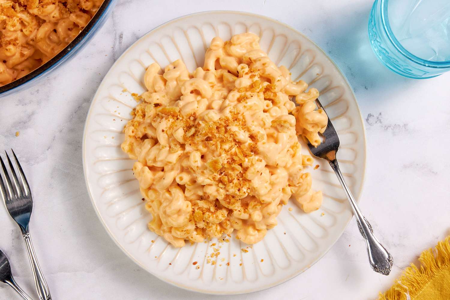 Plateful of one pot mac and cheese with a fork, and in the surroundings, a dutch oven with more, a glass of water, utensils, and a mustard kitchen towel