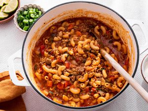 One-Pot Taco Pasta in a Pot, and in the Surroundings, a Bowl With Lime Wedges, a Bowl With Sliced Green Onions, a Bowl With Sour Cream, and a Mustard Colored Table Napkin