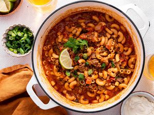 One-Pot Taco Pasta in a Pot, and in the Surroundings, a Bowl With Lime Wedges, a Bowl With Sliced Green Onions, a Bowl With Sour Cream, Glasses With a Drink, and a Mustard Colored Table Napkin