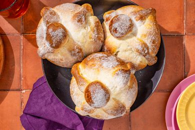 Pan de muerto in a bowl next to a stack of plates, a purple kitchen towel, and glasses of water, all on a brick surface
