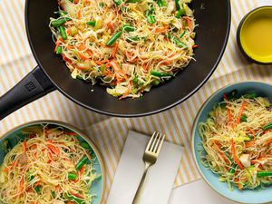 Pancit Bihon (Filipino Rice Noodles) in a Wok and Two Servings in Two Bowls at a Table Setting with Utensils and an Empty Bowl