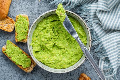 Table Knife With a Bit of Pea Pesto Rested on Top of a Bowl of Pea Pesto Surrounded by Slices of Toasted Bread on the Counter (Some Pieces of Bread Is Smeared With Pea Pesto) and a Blue and White Stripped Kitchen Towel 