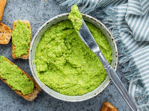 Table Knife With a Bit of Pea Pesto Rested on Top of a Bowl of Pea Pesto Surrounded by Slices of Toasted Bread on the Counter (Some Pieces of Bread Is Smeared With Pea Pesto) and a Blue and White Stripped Kitchen Towel 