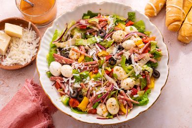 Bowl of Pizza Salad Surrounded by a Bowl of Grated Cheese With Blocks of Parmesan, a Cup With More Dressing, Breadsticks on the Counter, and a Pink Kitchen Towel