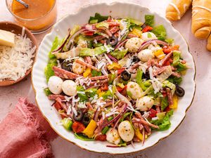 Bowl of Pizza Salad Surrounded by a Bowl of Grated Cheese With Blocks of Parmesan, a Cup With More Dressing, Breadsticks on the Counter, and a Pink Kitchen Towel