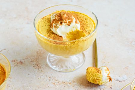 2-ingredient pumpkin mousse topped with whipped cream and a sprinkle of pumpkin spice, all in a sherbert glass. On the counter, a spoonful of mousse.