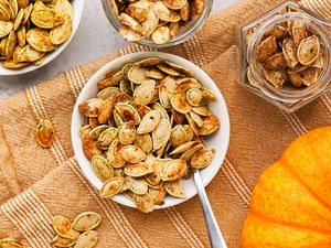 Bowl of toasted pumpkin seeds with a utensil, and in the surroudings, more toasted pumpkin seeds (with different toppings) in bowls and jars and an uncut pumpkin, all on light orange kitchen towel