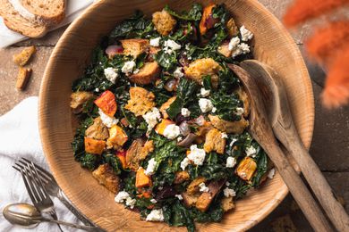 Bowl of Roasted Root Vegetable Panzanella Surrounded by a Slices of Bread, Vase of Flowers, Utensils, and a Kitchen Linen