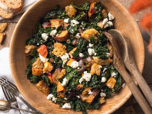 Bowl of Roasted Root Vegetable Panzanella Surrounded by a Slices of Bread, Vase of Flowers, Utensils, and a Kitchen Linen