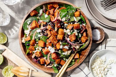 Bowl of Roasted Sweet Potato Salad Topped with Crumbled Feta Cheese, Surrounded by Stack of Plate with Forks, a Small Bowl of More Feta Cheese, a Small Bowl of Salt, a Cutting Board with Lime Wedges, a Kitchen Towel, and Empty Glasses