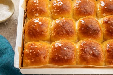 Salted honey rolls in a parchment paper-lined baking sheet on a cooling rack next to a blue kitchen towel and a bowl of flaky salt