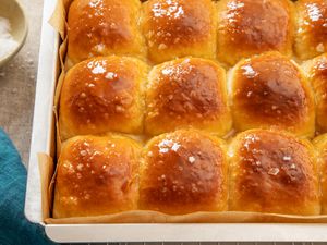 Salted honey rolls in a parchment paper-lined baking sheet on a cooling rack next to a blue kitchen towel and a bowl of flaky salt