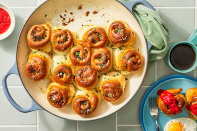 Savory breakfast rolls in a dutch oven pan at a table a setting with a stack of plates (top plate has a serving of rolls and eggs), a cup of coffee, and a small bowl of marinara sauce