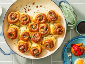 Savory breakfast rolls in a dutch oven pan at a table a setting with a stack of plates (top plate has a serving of rolls and eggs), a cup of coffee, and a small bowl of marinara sauce