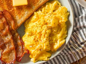 Photo of a plate of scrambled eggs, butter on toast, and slices of bacon on a white and gray kitchen towel with yellow lined illustration in the corner