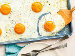 Wooden Spatula Lifting a Sheet Pan Fried Egg From a Baking Pan, and Next to the Sheet Pan, a Stack of Plate With Utensils and a Kitchen Towel