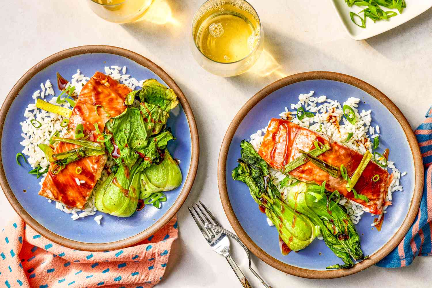 Two plates of 5-ingredient sheet pan salmon with rice and bok choy at a table setting with glasses of wine, small platter with sliced green onions, a couple of blue and pink table napkins, and utensils