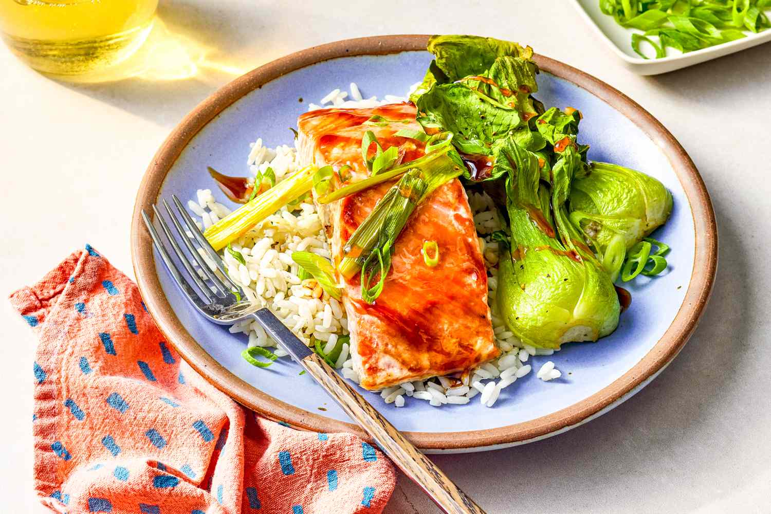 Plate of 5-ingredient sheet pan salmon with rice and bok choy at a table setting with glasses of wine, small platter with sliced green onions, and a blue and pink table napkin