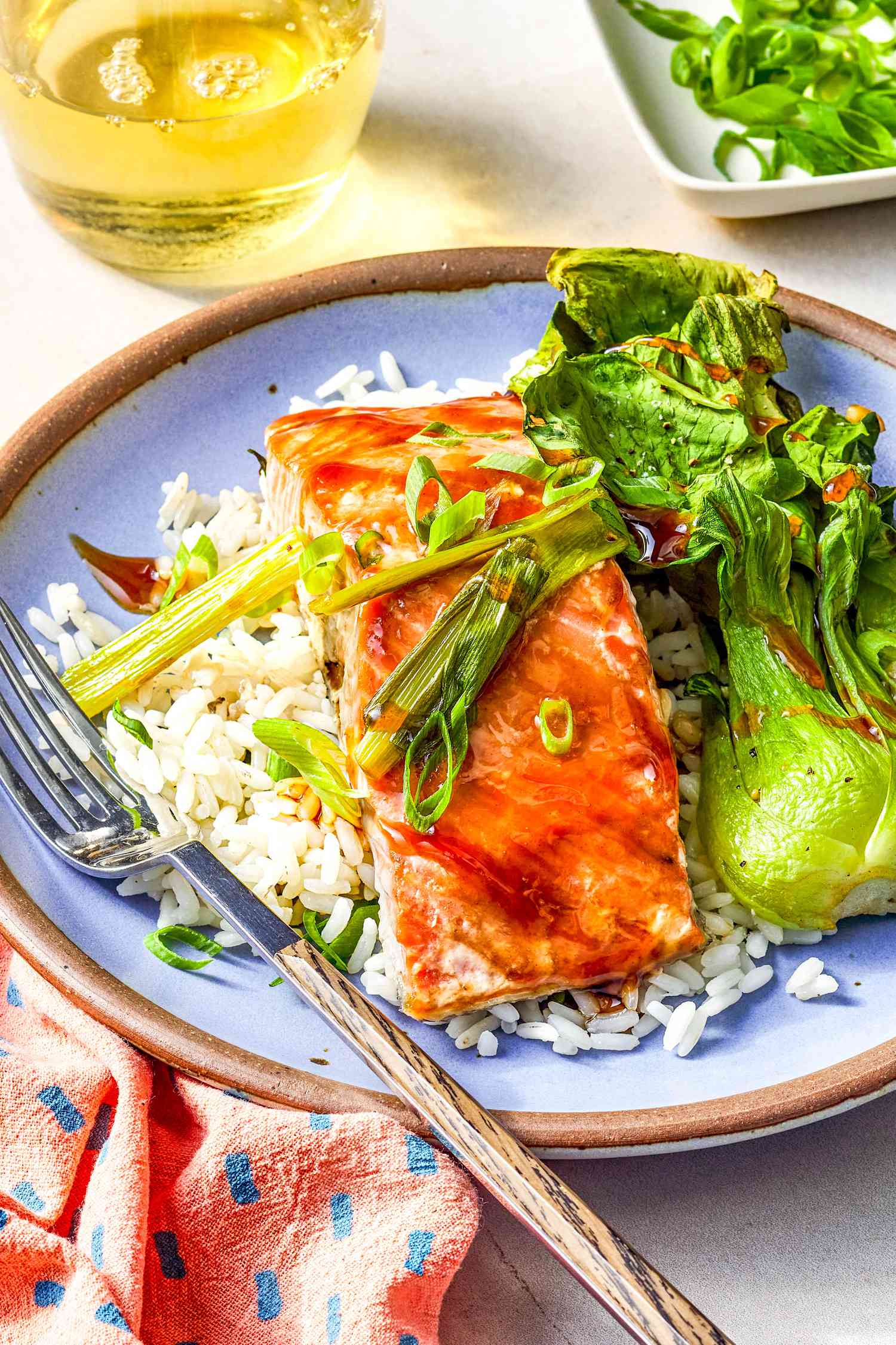 Plate of 5-ingredient sheet pan salmon with rice and bok choy at a table setting with glasses of wine, small platter with sliced green onions, and a blue and pink table napkin