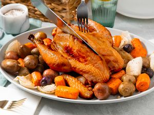 Carving knife cutting into simple roast chicken that's served on a bed of roasted vegetables at a table setting with plates, a bread basket, a glass, and utensils 