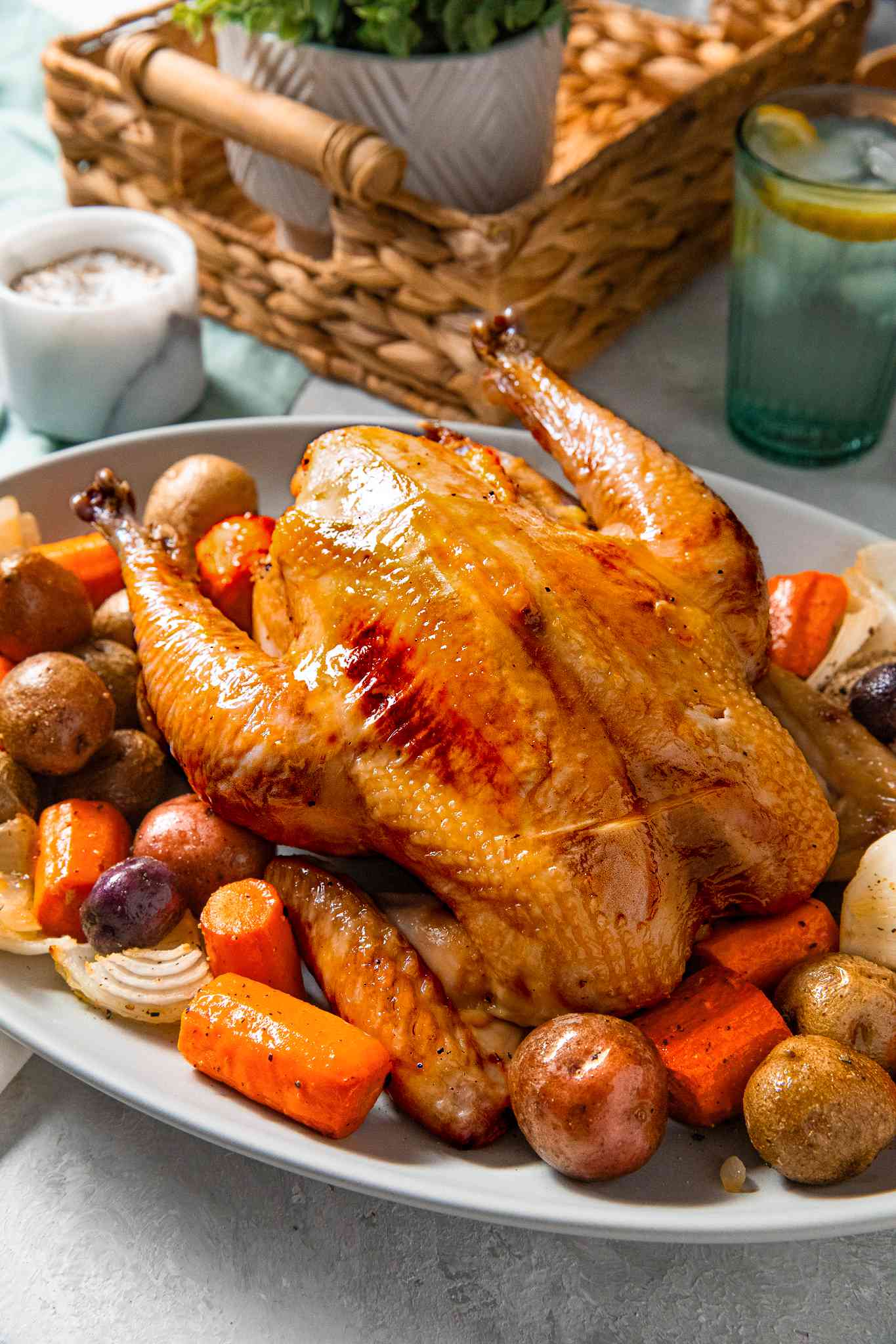 Simple roast chicken served on a bed of roasted vegetables at a table setting with jar of seasoning, a glass of lemon ice water, and a basket with a potted plant