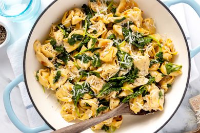 Skillet Cacio e Pepe Tortellini With Wilted Greens in a Pot With a Serving Spoon, All on a White Kitchen Towel on the Counter, and Next to It, a Bowl of Cracked Pepper, a Glass of Water, and a Block of Pecorino Romani Cheese 