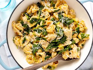 Skillet Cacio e Pepe Tortellini With Wilted Greens in a Pot With a Serving Spoon, All on a White Kitchen Towel on the Counter, and Next to It, a Bowl of Cracked Pepper, a Glass of Water, and a Block of Pecorino Romani Cheese 