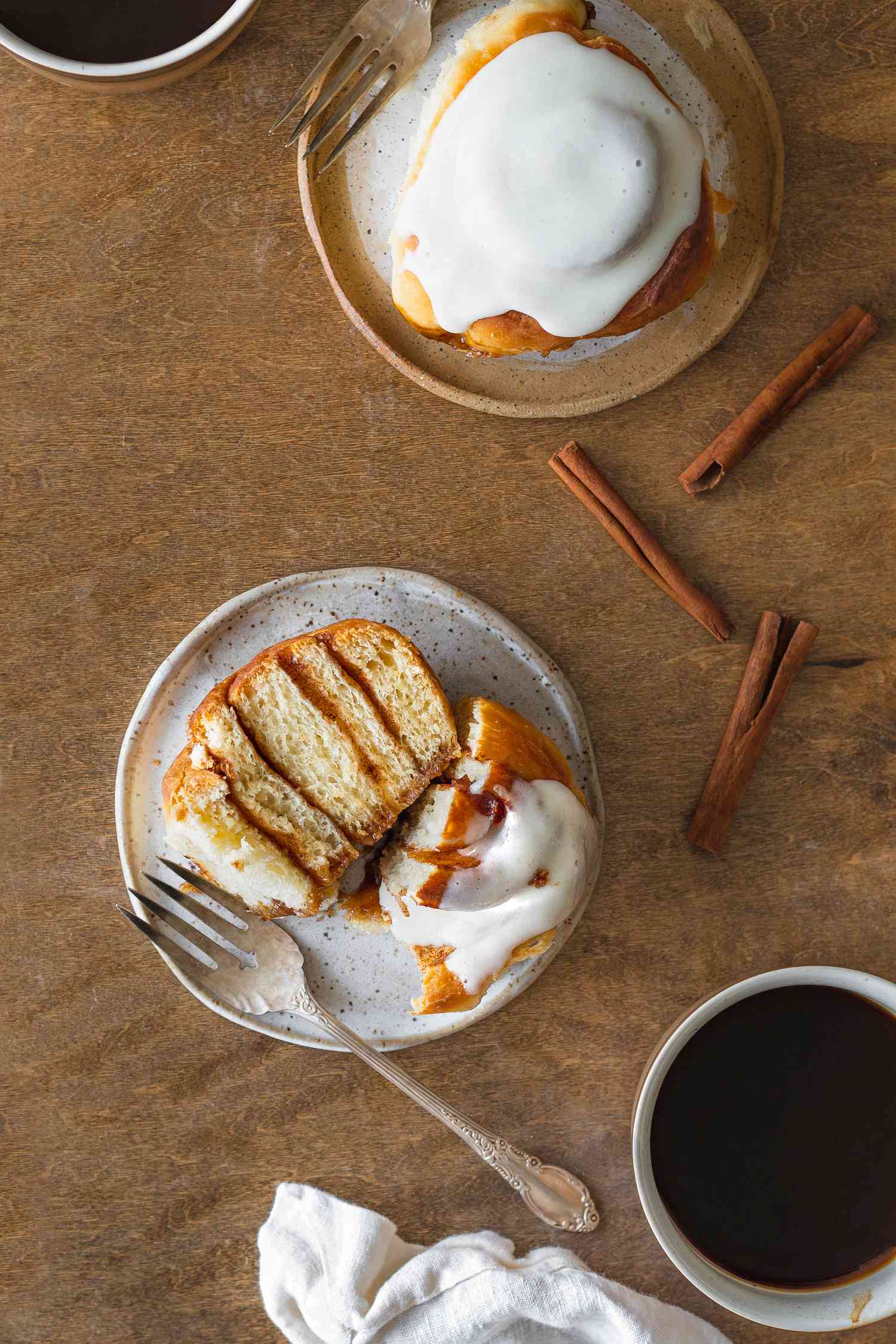 Sourdough Discard Cinnamon Roll on a Small Plate Cut in Half Surrounded by a Cup of Coffee, Cinnamon Sticks, and Another Cinnamon Roll on a Plate