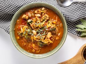 Spaghetti Squash Soup with Italian Sausage in a Bowl Next to a Kitchen Towel, a Spoon, Fresh Sage Leaves, and a Wooden Holder with a Grater