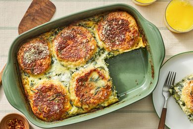 Spinach florentine breakfast casserole in a casserole dish at a table setting with a serving on a plate, glasses of orange juice, a bowl of crushed pepper, and a spatula