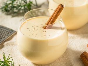 Glass of eggnog with a cinnamon stick in it, and in the surroundings, another glass of eggnog, cinnamon sticks, a handheld grater, and rosemary 