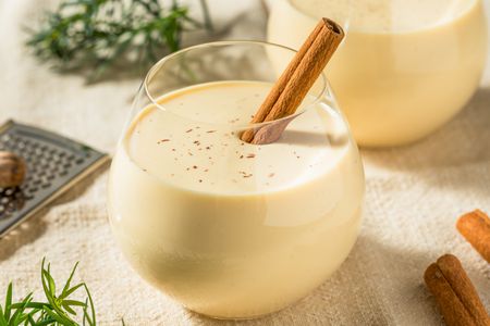 Glass of eggnog with a cinnamon stick in it, and in the surroundings, another glass of eggnog, cinnamon sticks, a handheld grater, and rosemary 