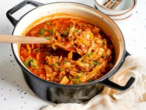 Sweet and Sour Cabbage Soup in a Large Pot with Some Raised Up Using a Wooden Spoon, and Surrounding the Pot, a Kitchen Linen and a Stack of Bowls with Utensils