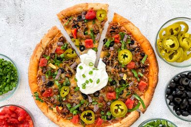 Taco Pizza on the Counter With a Slice Slightly Removed from the Pizza, and in the Surroundings, Bowls of Toppings (L to R: Sliced Scallions, Salsa, Sliced Lettuce, Sliced Olives, and Sliced JalapeÃ±os)