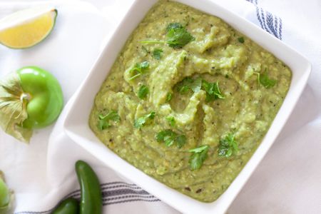 Bowl of Tomatillo Avocado Salsa Surrounded by Tomatillos, Lime Wedges, and Jalapeños