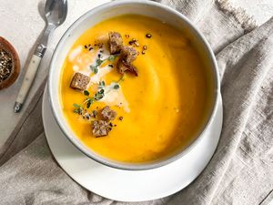 Bowl of Vegan Butternut Squash Soup Topped with Thyme, Coconut Milk, Croutons, and Cracked Pepper, Sitting on a Kitchen Linen and Surrounded by a Bowl of Cracked Pepper and a Spoon 