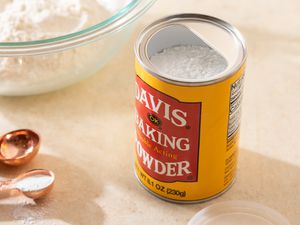 Can of baking powder surrounded by tablespoons with some baking powder in one of the spoons and a bowl of flour