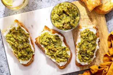 Slices of Toast Topped With Ricotta and Zucchini Butter on a Marble Slab Next to a Bowl With More Zucchini Butter and a Spoon, and in the Surroundings, a Mustard Colored Kitchen Towel and a Glass