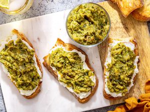 Slices of Toast Topped With Ricotta and Zucchini Butter on a Marble Slab Next to a Bowl With More Zucchini Butter and a Spoon, and in the Surroundings, a Mustard Colored Kitchen Towel and a Glass