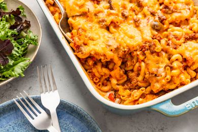 Johnny Marzetti casserole in a casserole dish, and in the surroundings, a plate with two forks and a bowl of mixed greens salad