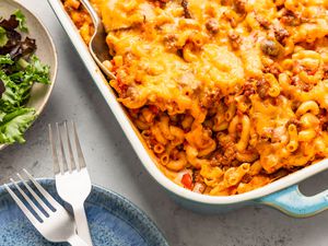 Johnny Marzetti casserole in a casserole dish, and in the surroundings, a plate with two forks and a bowl of mixed greens salad