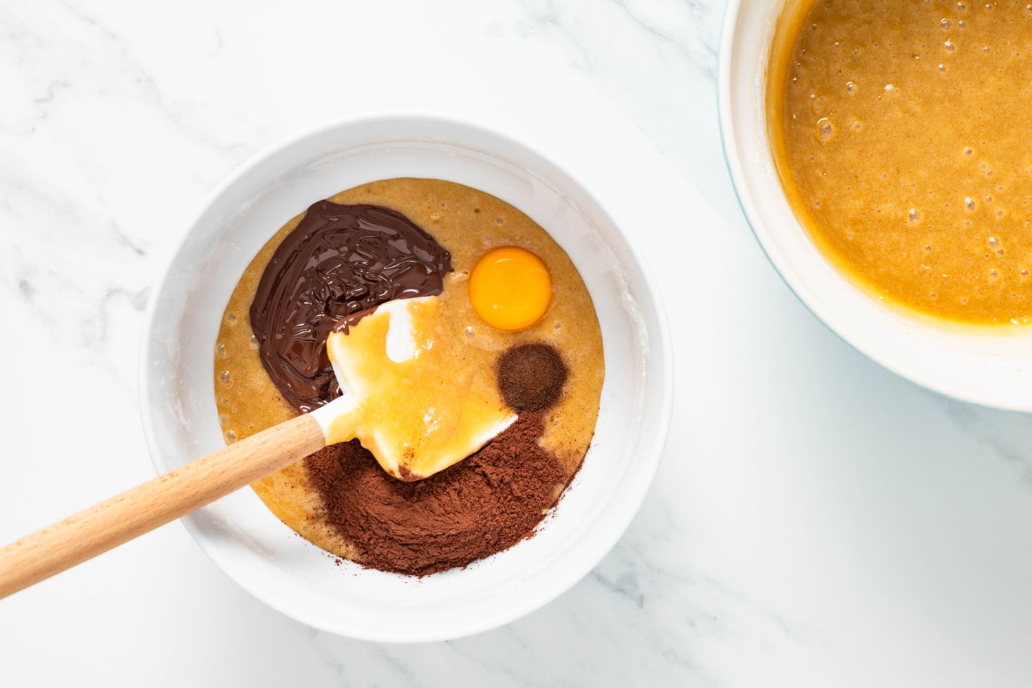 Bowl with some of the blondie batter, an egg yolk, melted chocolate, and Dutch processed cocoa powder next to the bowl with the blondie batter for marble brownies recipe