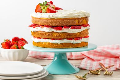 Strawberry Shortcake Cake on a Cake Stand, and in the Surroundings, a Stack of Plates, a Bowl With Whole Strawberries, a Kitchen Towel, and a Few Forks on the Counter
