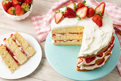 Strawberry Shortcake Cake Topped With Halved Strawberries on a Cake Stand, and on the Counter Next to It, a Plate With a Slice of Cake, a Bowl of Strawberries, and a Kitchen Towel