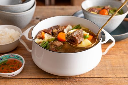 Ladle raising some nilagang baka from a Dutch oven with more, and in the surroundings, a stack of bowls, a serving of Filipino beef soup in a bowl, a bowl of rice, and a small bowl of fish sauce